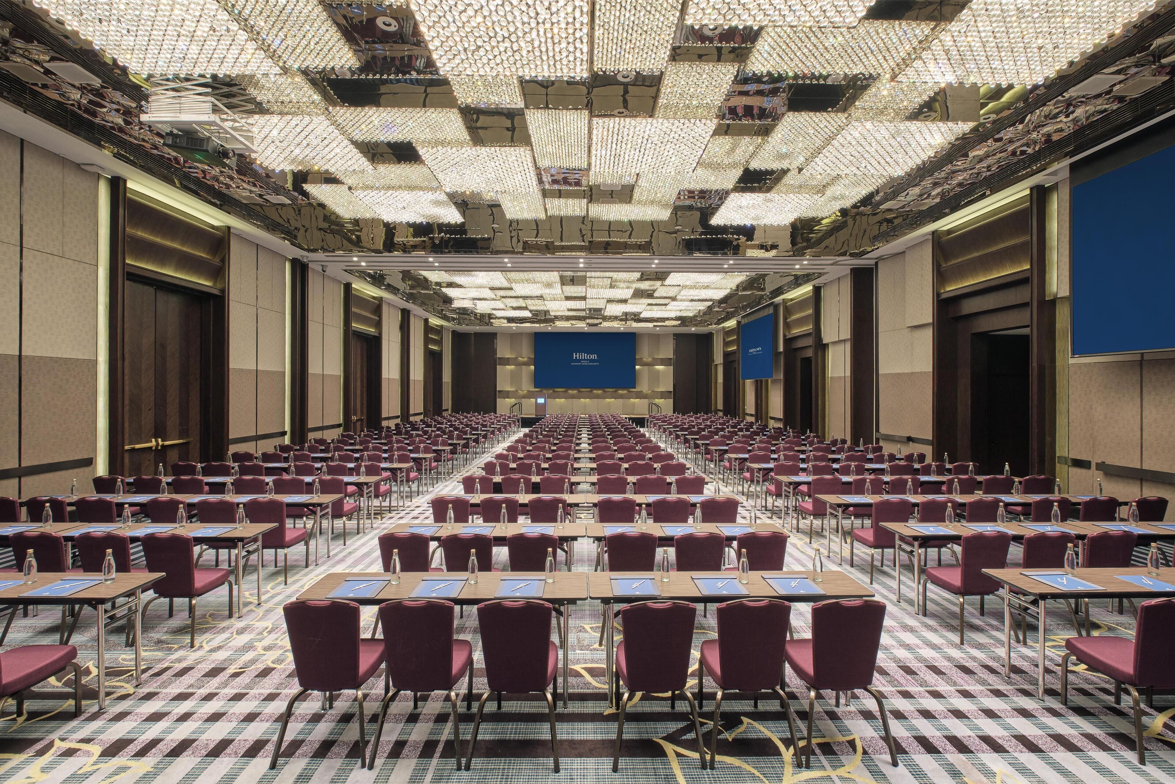 Un salón de conferencias amplio con filas de mesas y sillas, una alfombra estampada y un techo con una rejilla de luces.