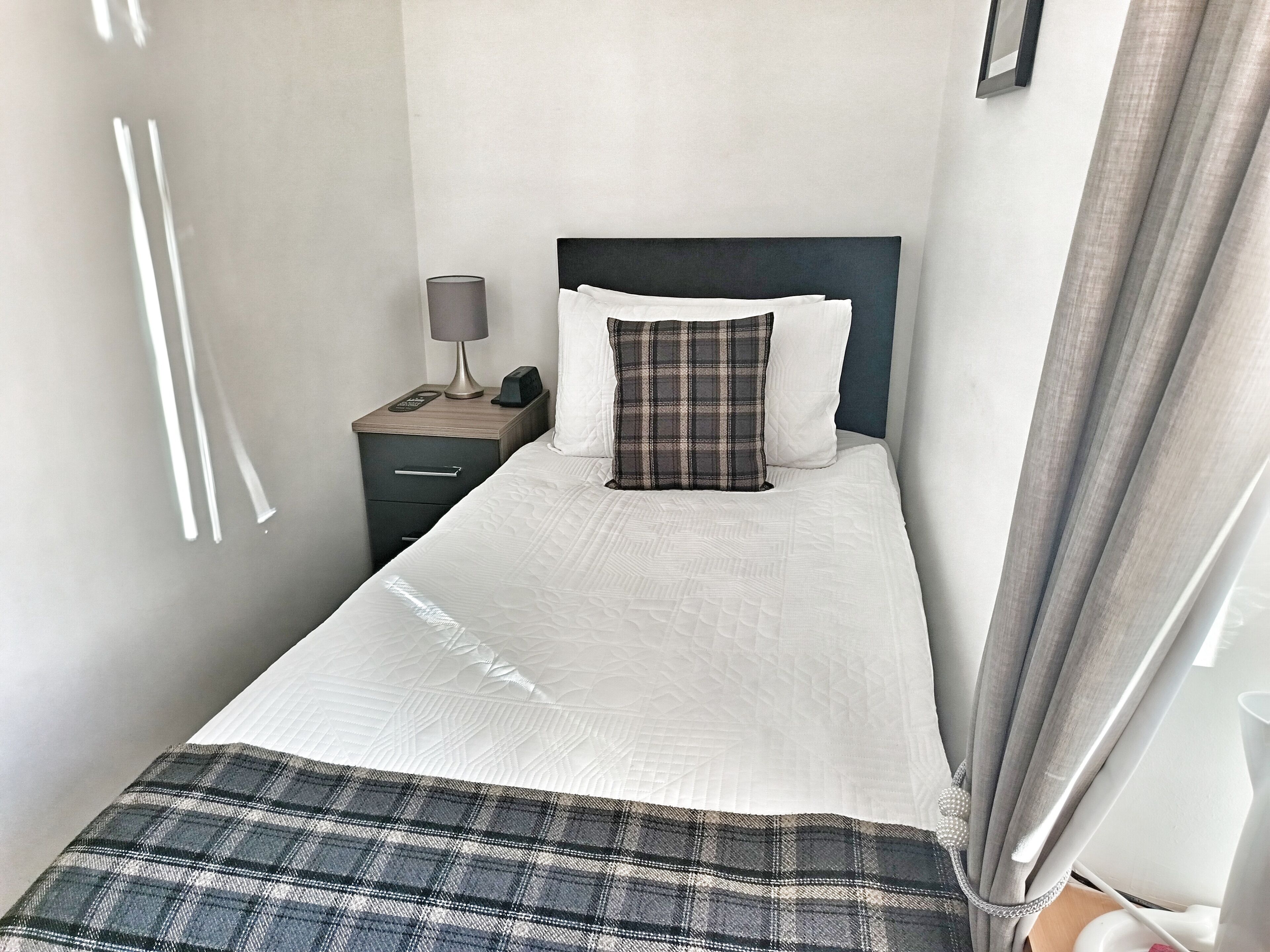 Una cama pequeña y bien hecha con edredón blanco y almohada de cuadros, una mesita de noche con lámpara, y una cortina parcialmente abierta.