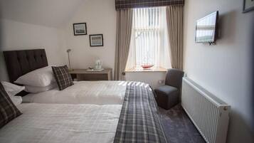 A bedroom with two beds, a desk with a chair, a radiator, and a window with curtains.