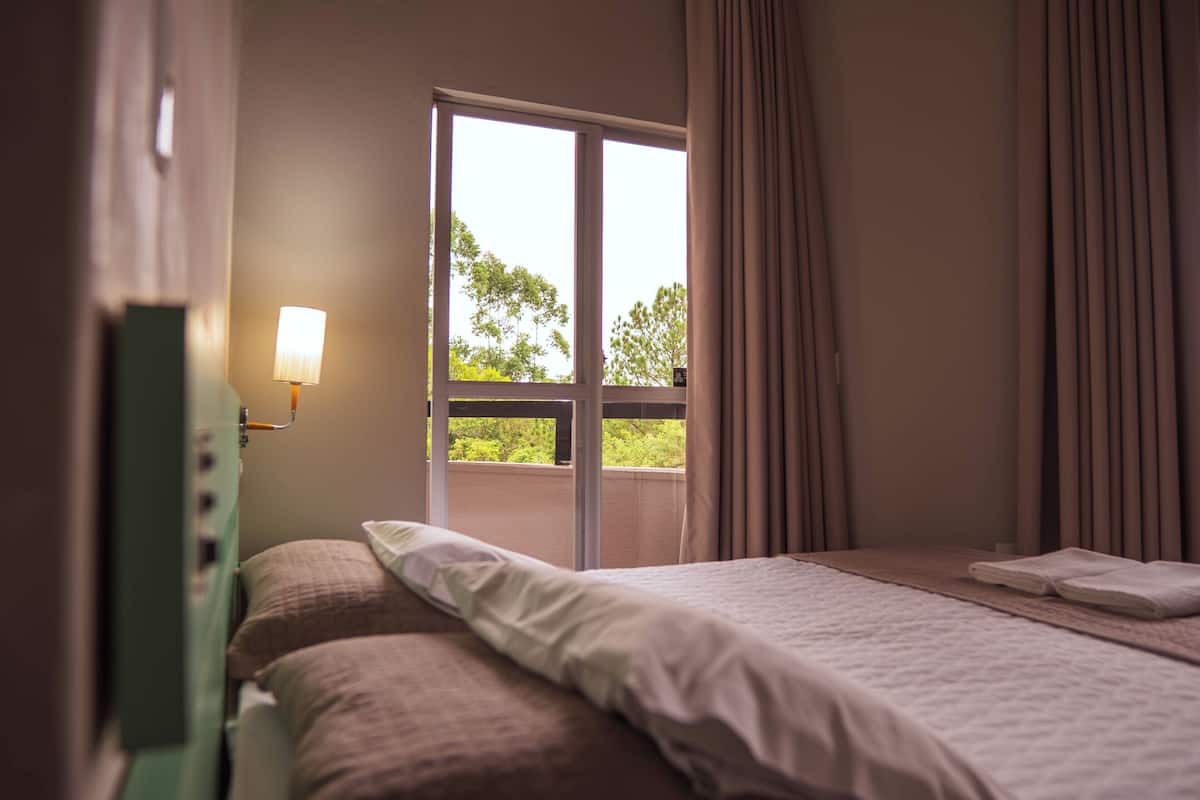 A hotel room with a bed, a nightstand with a lamp, a window with curtains, and a view of greenery outside.