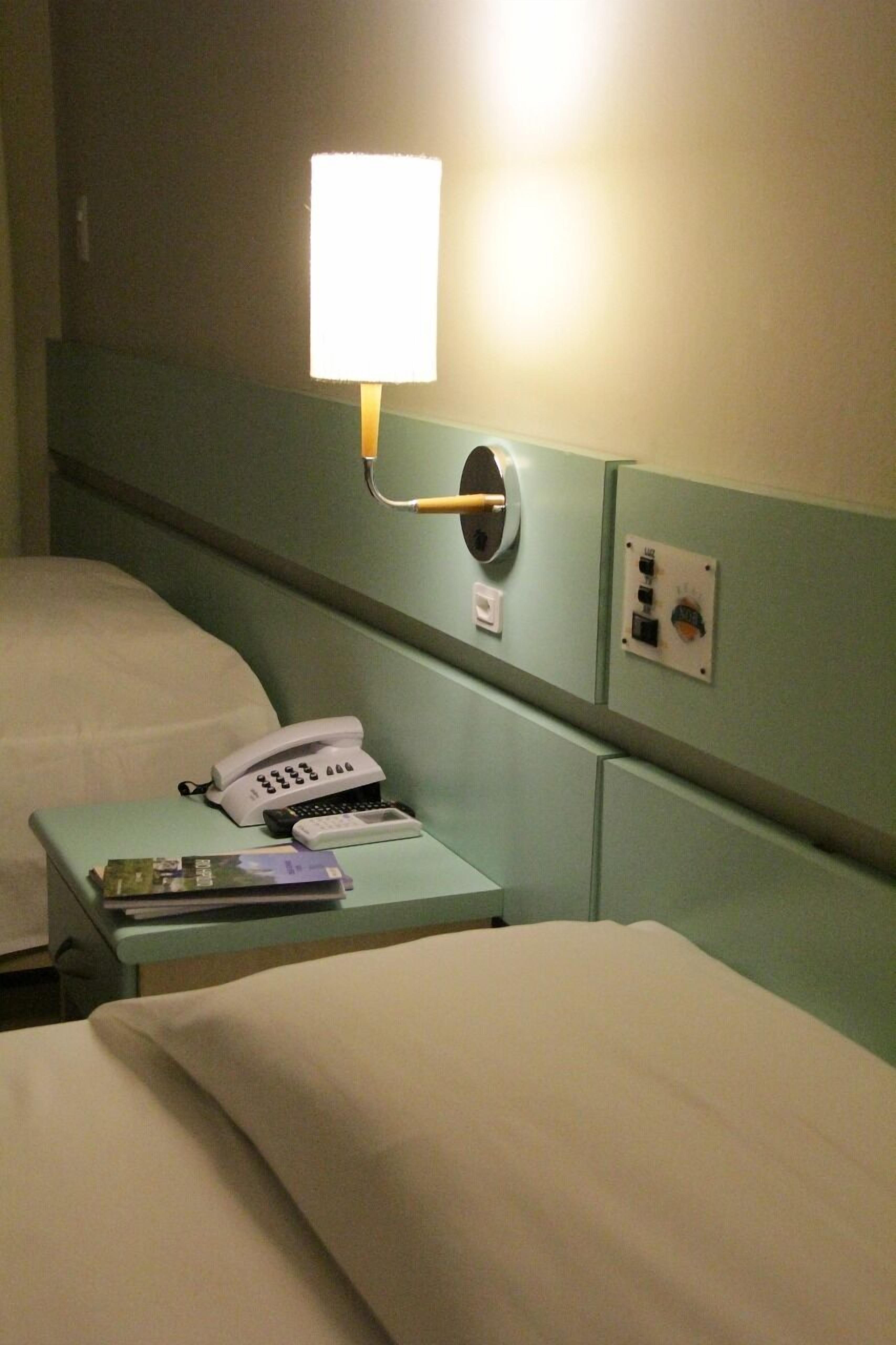 A hotel room with two beds, a bedside phone, a lamp, and a control panel on the wall.