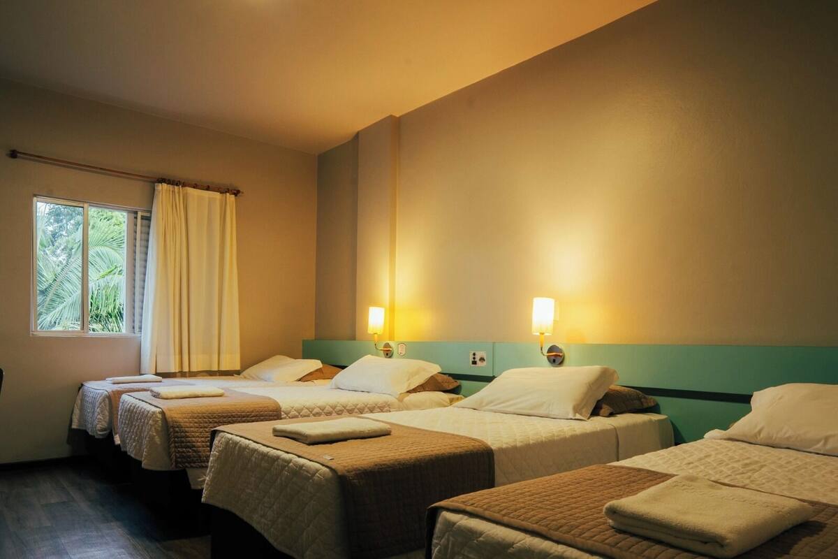 A hotel room with three beds, a window with curtains, and wall-mounted lamps.