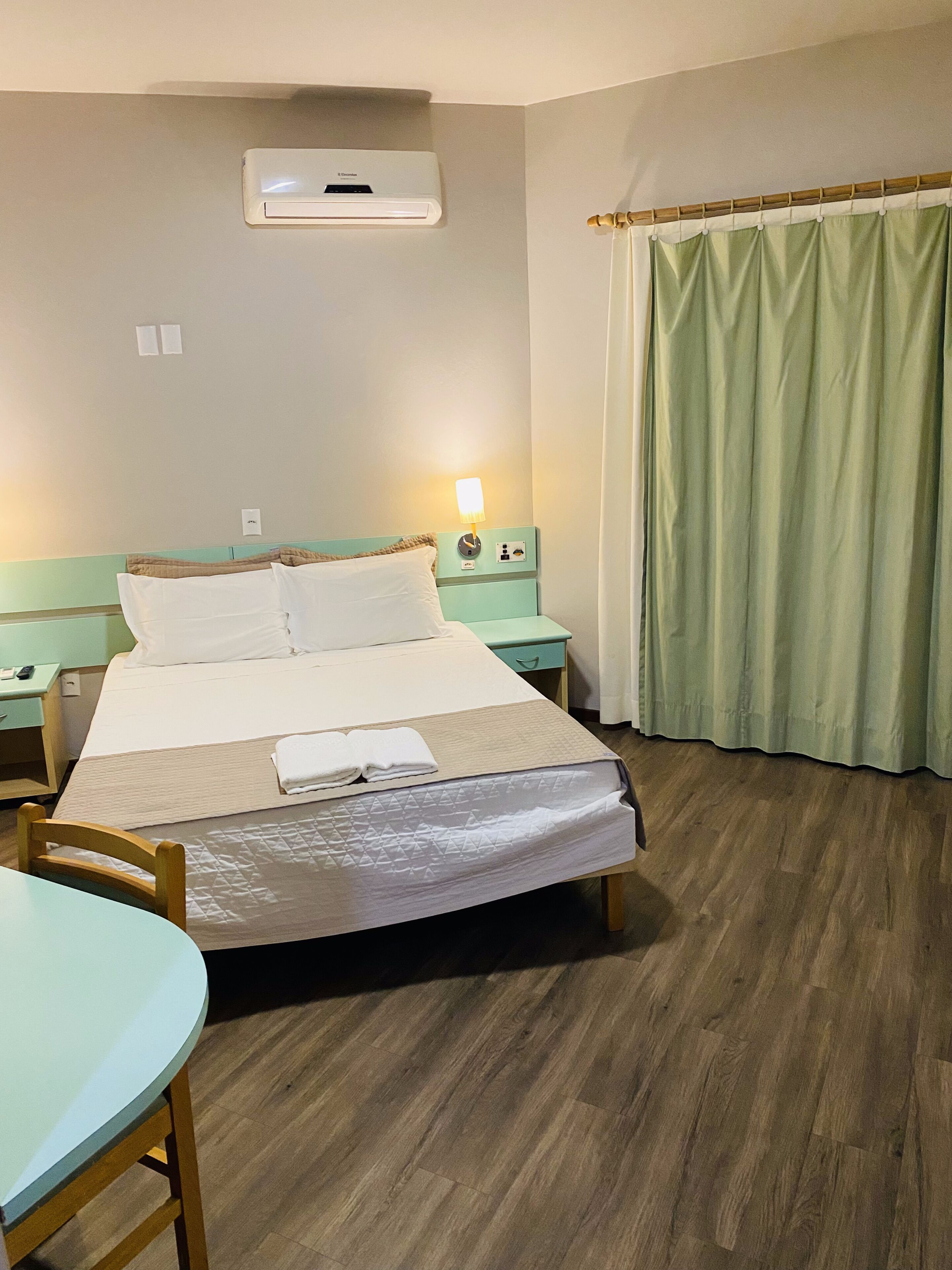 A hotel room with a bed, bedside tables, a lamp, a window with curtains, and an air conditioning unit.