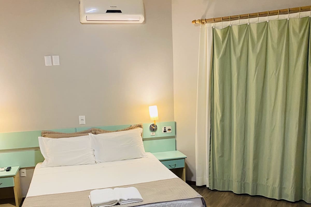 A hotel room with a bed, bedside tables, a lamp, a window with curtains, and an air conditioning unit.