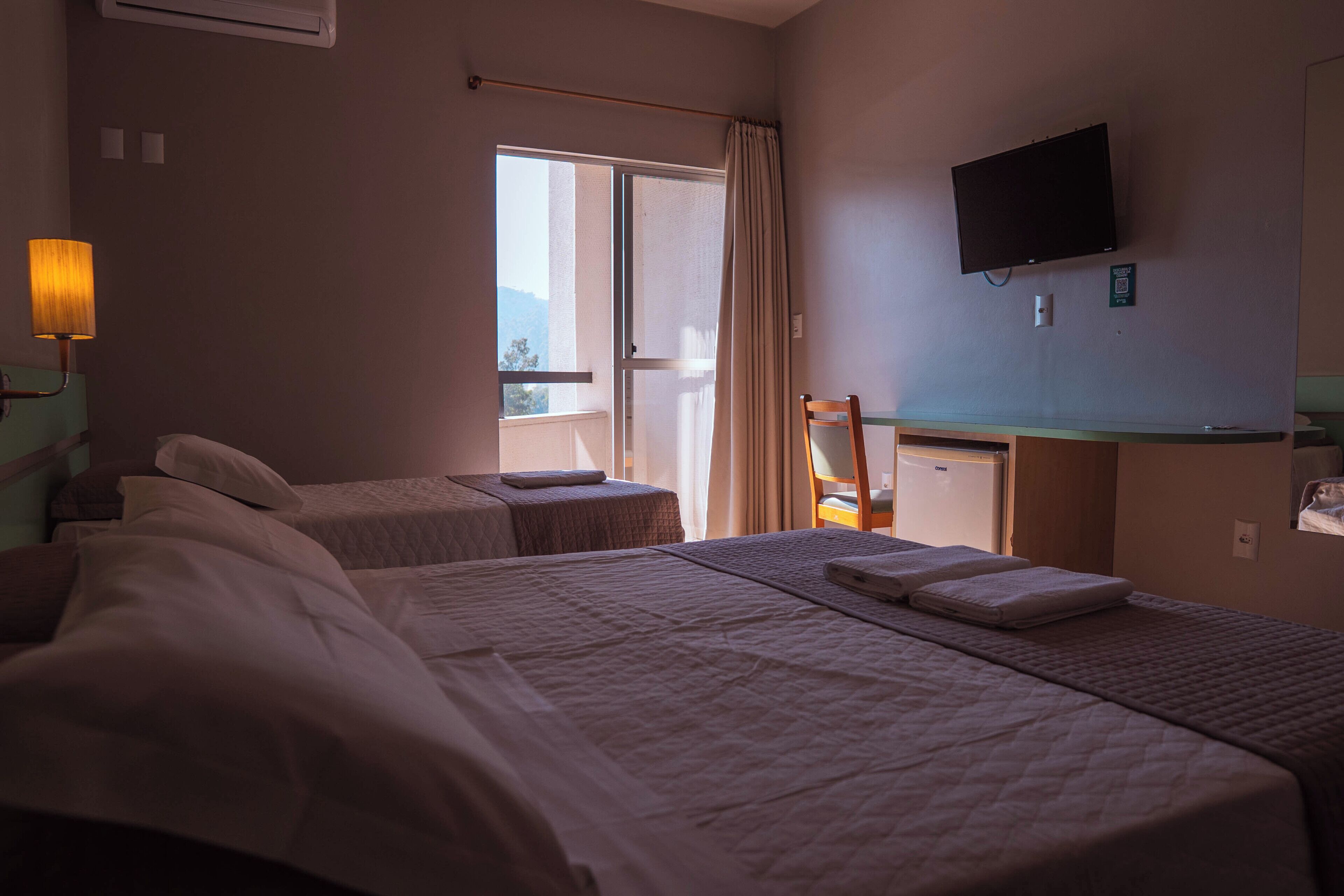A hotel room with two beds, a television, a desk, and a window with curtains.