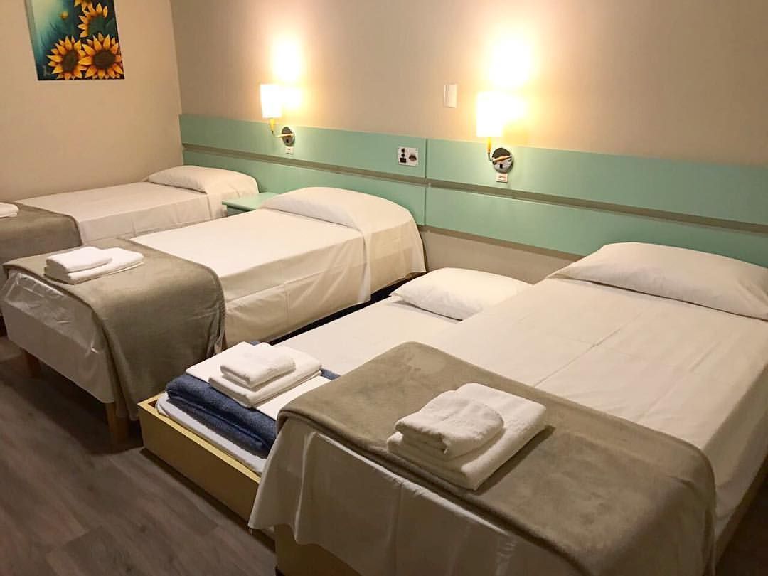 A hotel room with four single beds, each with a white bedspread and a folded towel. The room has a teal-colored headboard and wall-mounted lights.