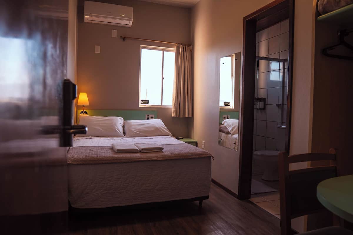 A hotel room with a bed, a bedside table, a lamp, a window with curtains, a mirror, and a bathroom visible through an open door.
