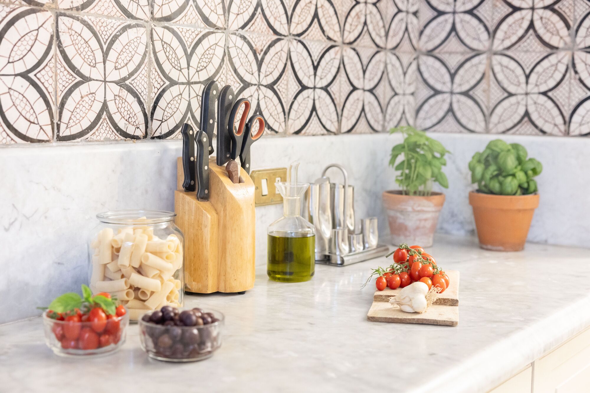 A kitchen counter with pasta, tomatoes, and a cutting board. There are also potted plants and a knife block.