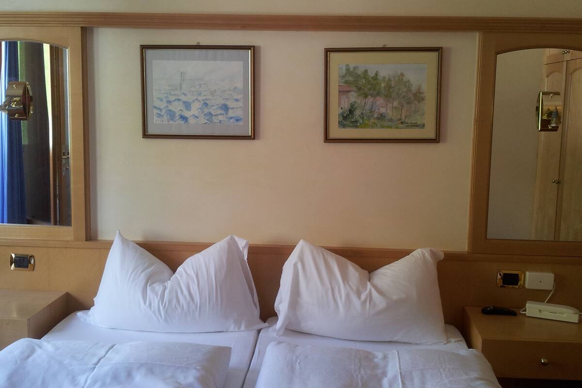 A hotel room with a bed, two framed paintings on the wall, a nightstand with a remote control, and a mirror.