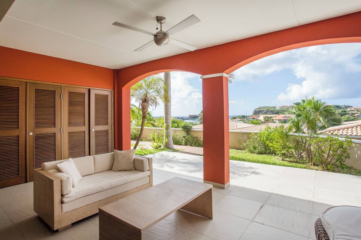 Amplia sala de estar con sofá, mesa de centro, ventilador de techo y vista a un paisaje exuberante con palmeras y una colina distante.