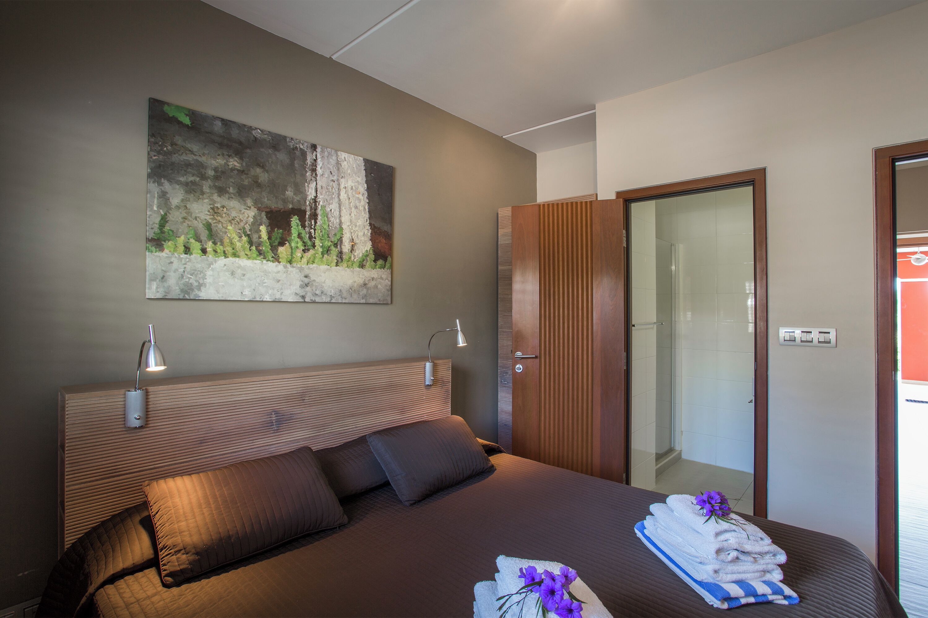 Ein Hotelzimmer mit Bett, einem Bild an der Wand, einer Tür und einem durch eine Glasscheibe einsehbaren Badezimmer.