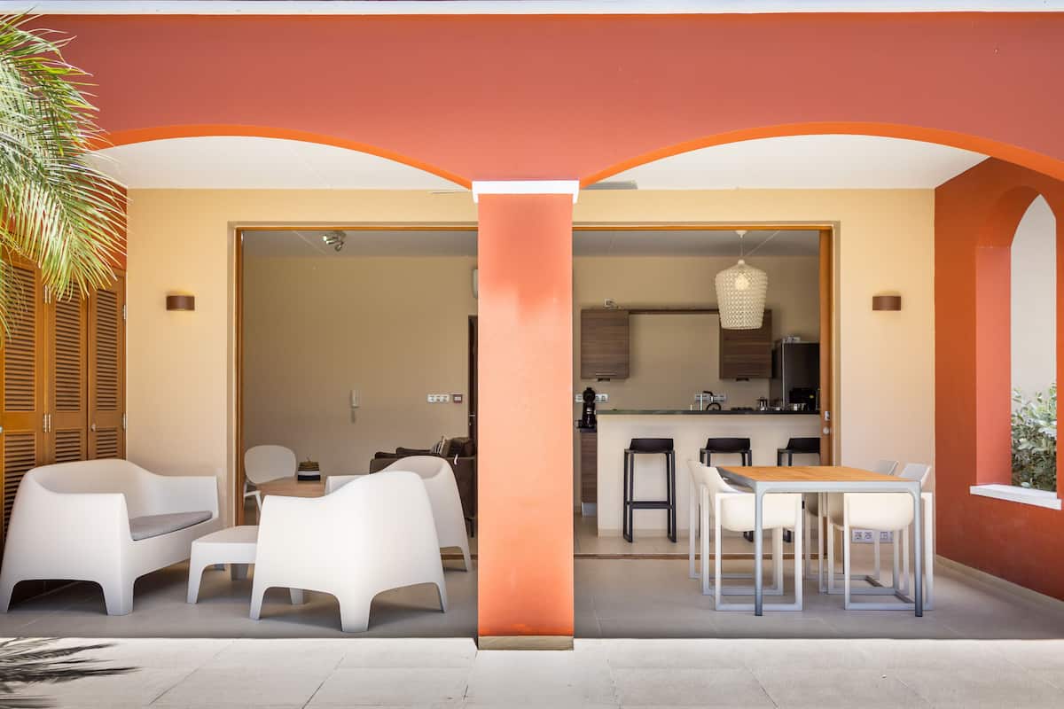 Un espacio exterior moderno para comer con sillas blancas y una mesa de madera, conectado a una cocina con barra y taburetes.