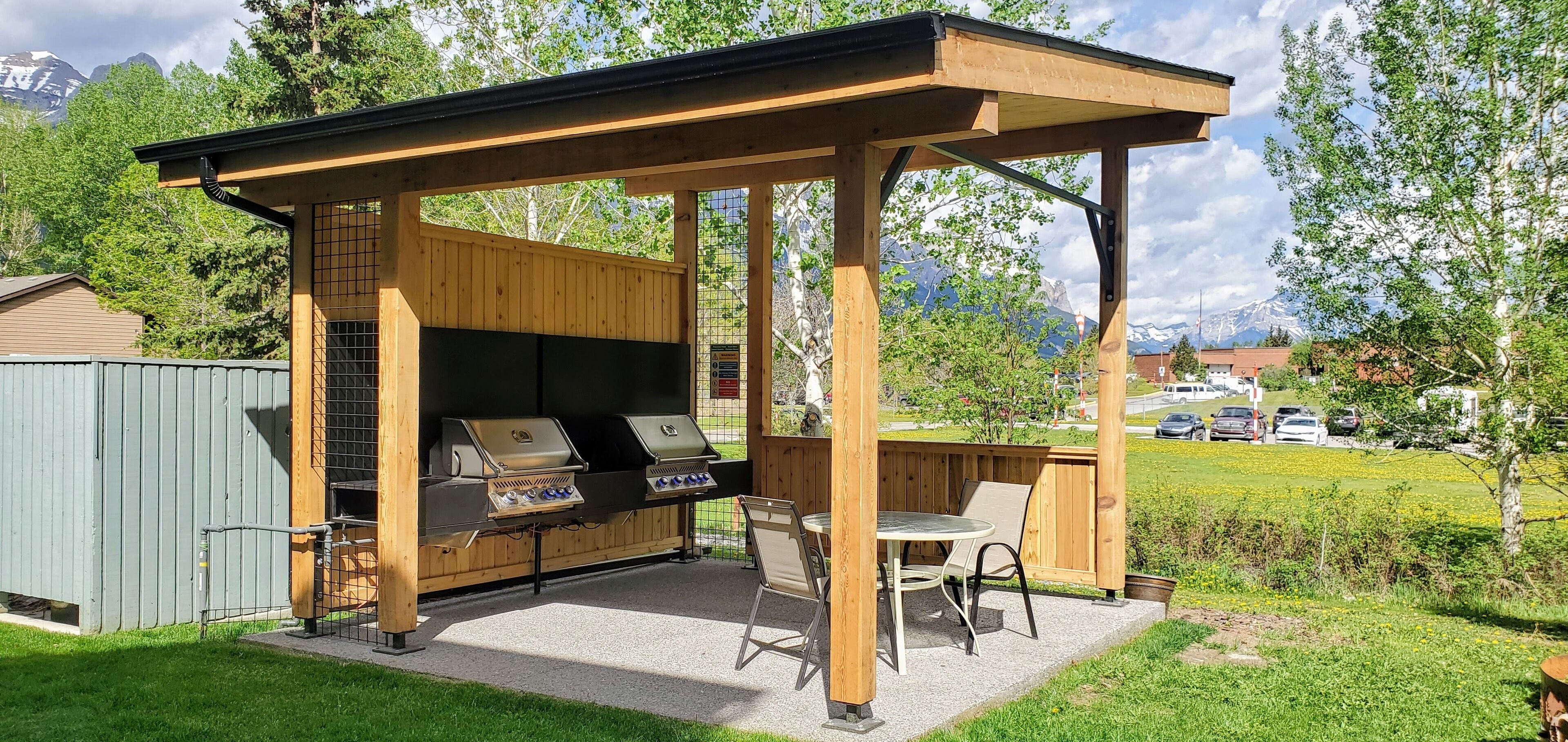 An outdoor barbecue area with a wooden pergola, two grills, and a table with chairs.