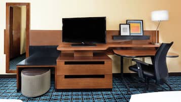 A hotel room with a wooden desk, a flat-screen TV, a chair, a small ottoman, and a lamp.
