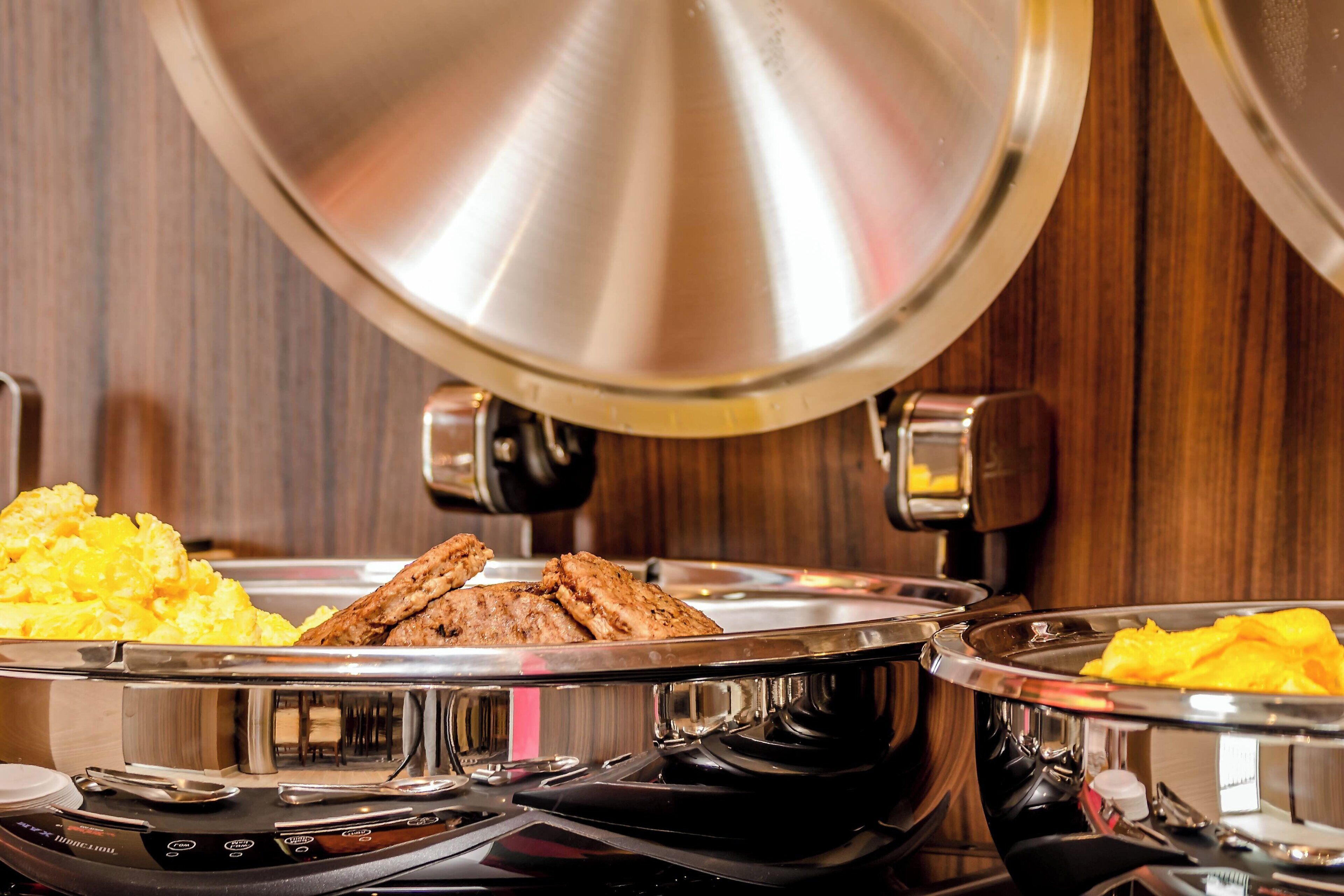 A buffet setup with various food items, including scrambled eggs and meat, on stainless steel trays.