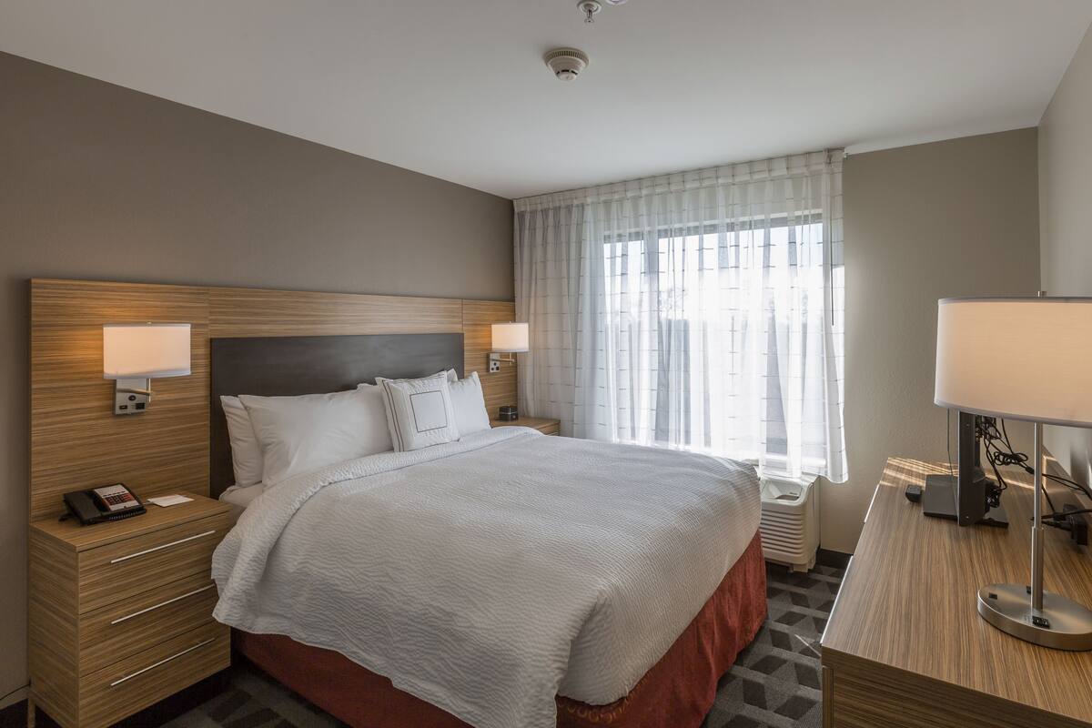 Ein modernes Hotelzimmer mit einem großen Bett, Nachttischlampen, einem Schreibtisch mit Lampe und einem Fenster mit durchsichtigen Vorhängen.