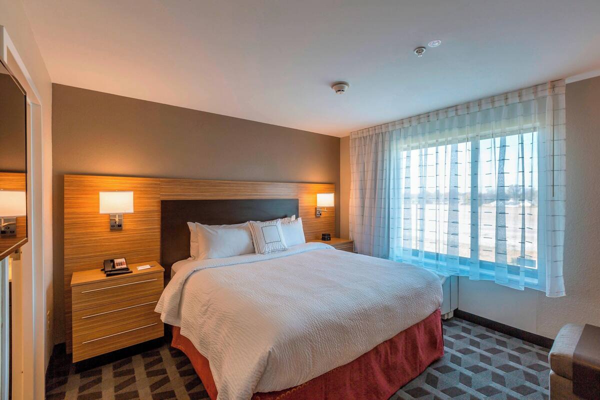 Ein Hotelzimmer mit einem großen Bett, zwei Nachttischen mit Lampen, einem Fenster mit durchsichtigen Vorhängen und einem gemusterten Boden.