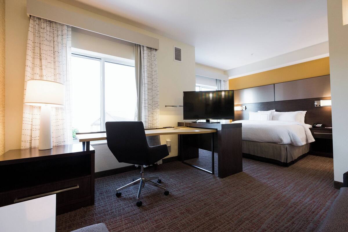 Una habitación de hotel con cama, escritorio, silla, televisor y ventana con cortinas.