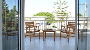 A balcony with two wooden chairs and a small table, a glass door, and a view of a tree and buildings. - Hotel Anna Apartments (Gouves)