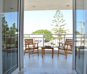 Un balcon avec deux chaises en bois et une petite table, une porte vitrée et une vue sur un arbre et des immeubles.