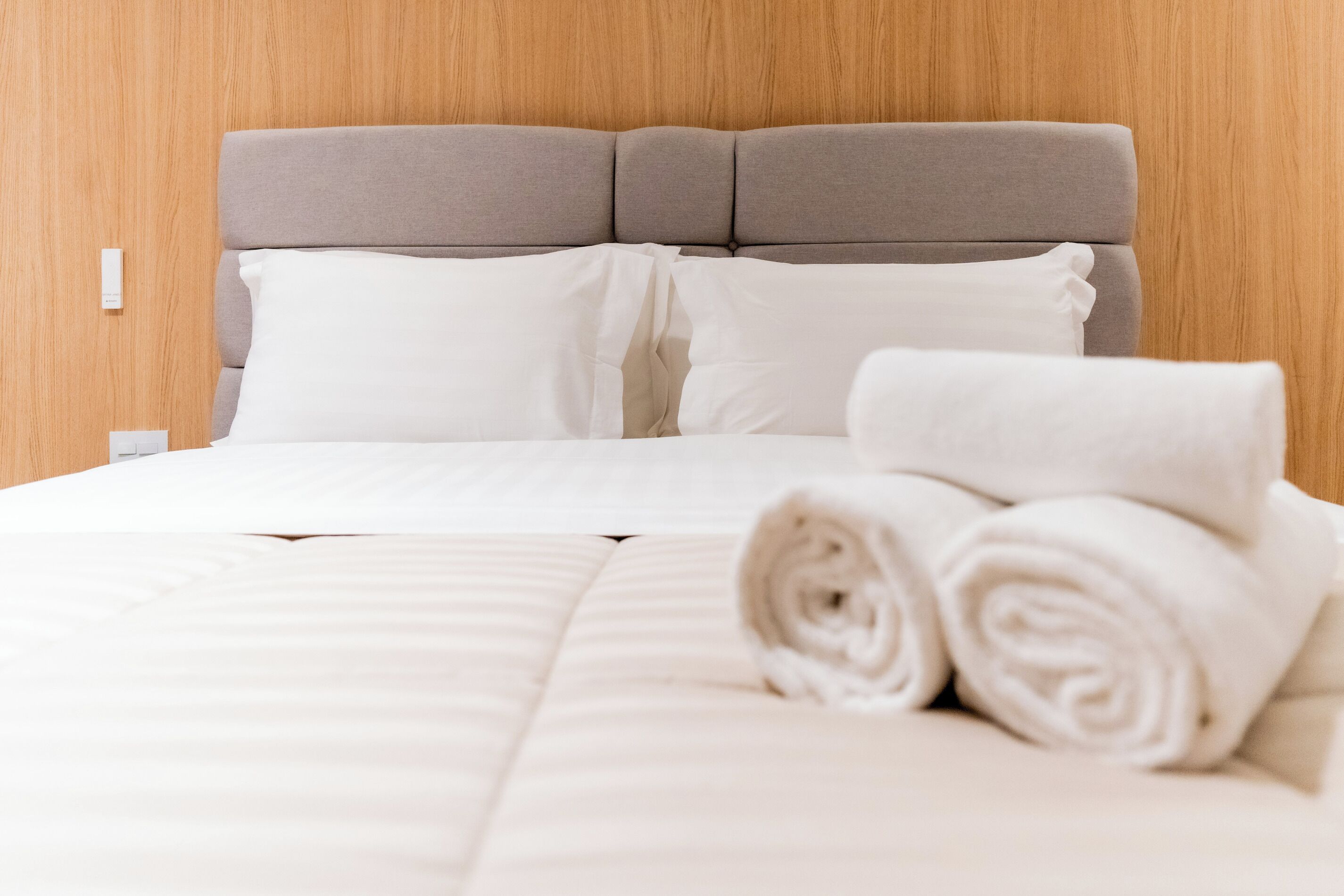 A neatly made bed with white linens and pillows, a grey headboard, and a wooden wall.