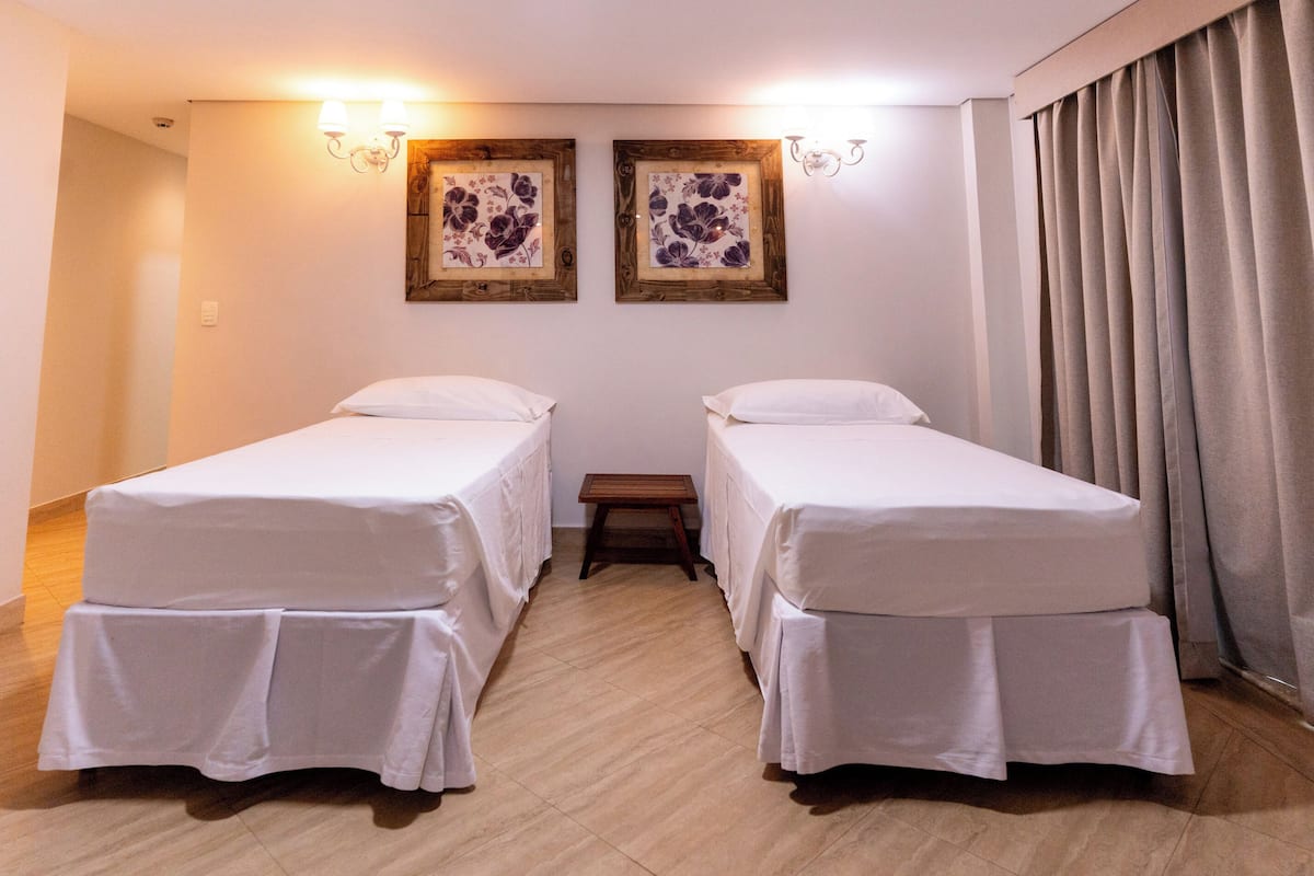 A hotel room with two single beds, a small wooden stool, and two wall-mounted light fixtures.