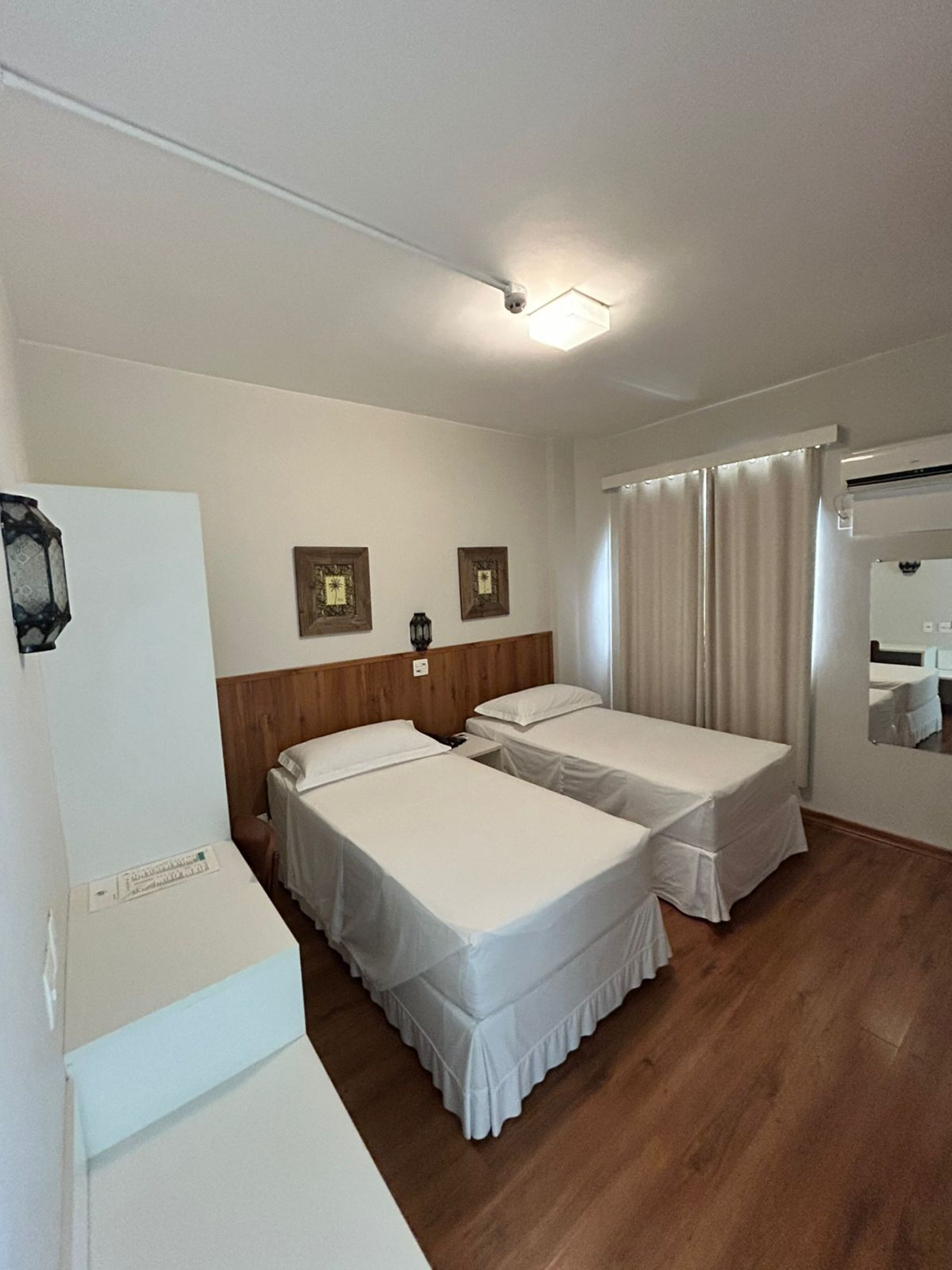 A hotel room with two single beds, a wooden headboard, a small table, a mirror, and a ceiling light.