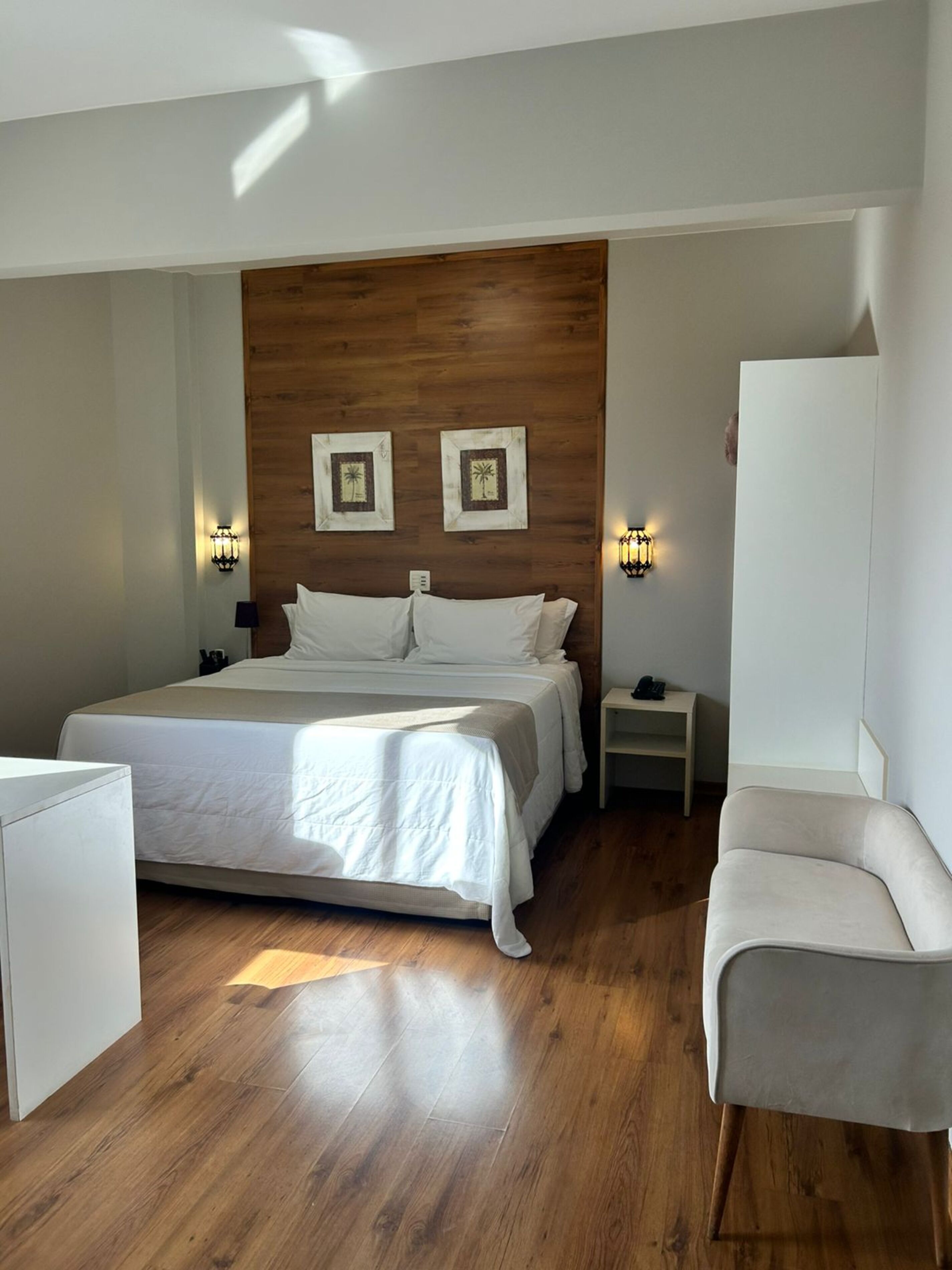 Un dormitorio con cabecera de madera, cama con ropa blanca, dos luces fijadas a la pared, una mesita de noche con un teléfono y un sillón gris.