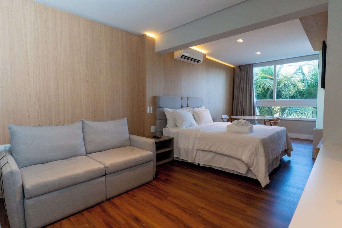 A modern hotel room with a wooden floor, a sofa, a bed with white linens, and a window with a view of palm trees.