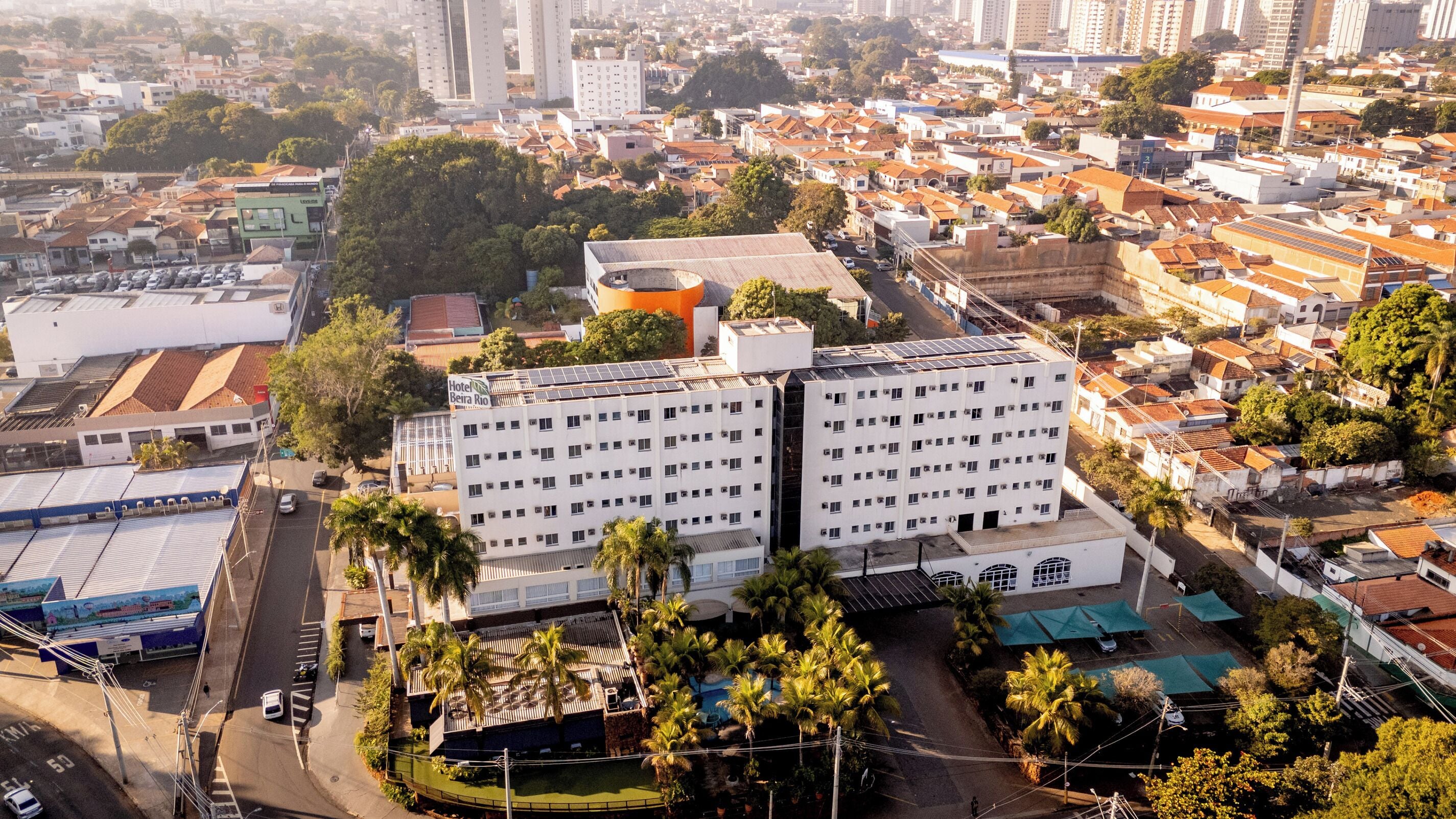 Um hotel com vários andares, com um jardim no terraço e painéis solares, situado numa área urbana densa com diversos edifícios e vegetação.