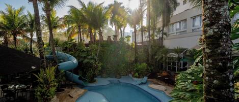 A pool area with a slide, surrounded by palm trees and plants, and a building in the background.