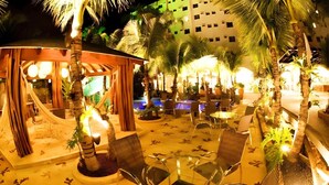 An outdoor lounge area with seating and a pool, surrounded by palm trees and a building in the background. - Beira Rio Palace Hotel (Piracicaba)