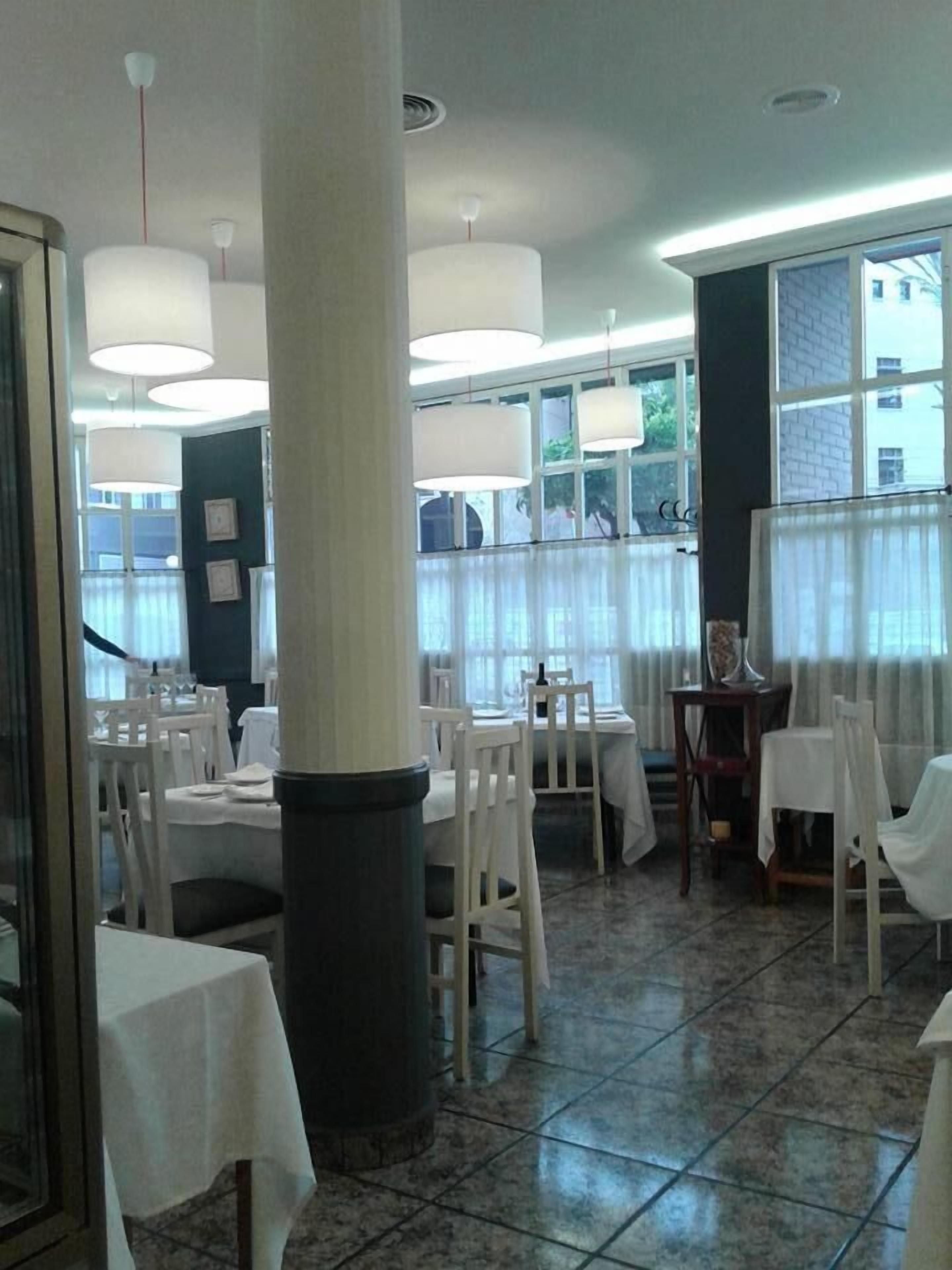 A dining area with white tables and chairs, large windows with curtains, and hanging pendant lights.