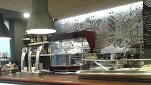A cafe with a counter, two stools, a coffee machine, and a decorative tiled wall. - Hotel Los Bartolos (Alhama de Murcia)
