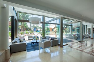 A spacious lobby with a modern design, featuring a seating area with sofas and armchairs, a coffee table, and large windows offering a view of the outdoor garden and palm trees.