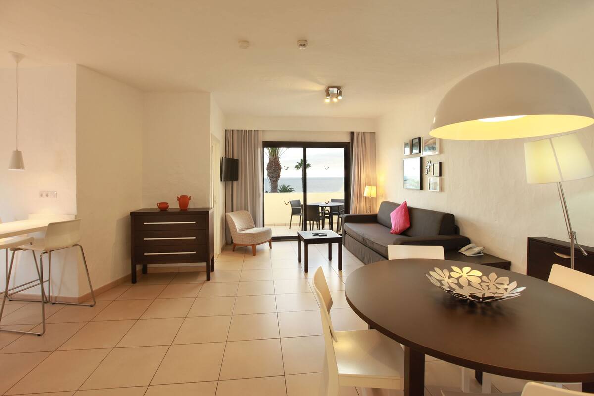 A hotel room with a dining area, a sofa, a TV, and a view of the beach.
