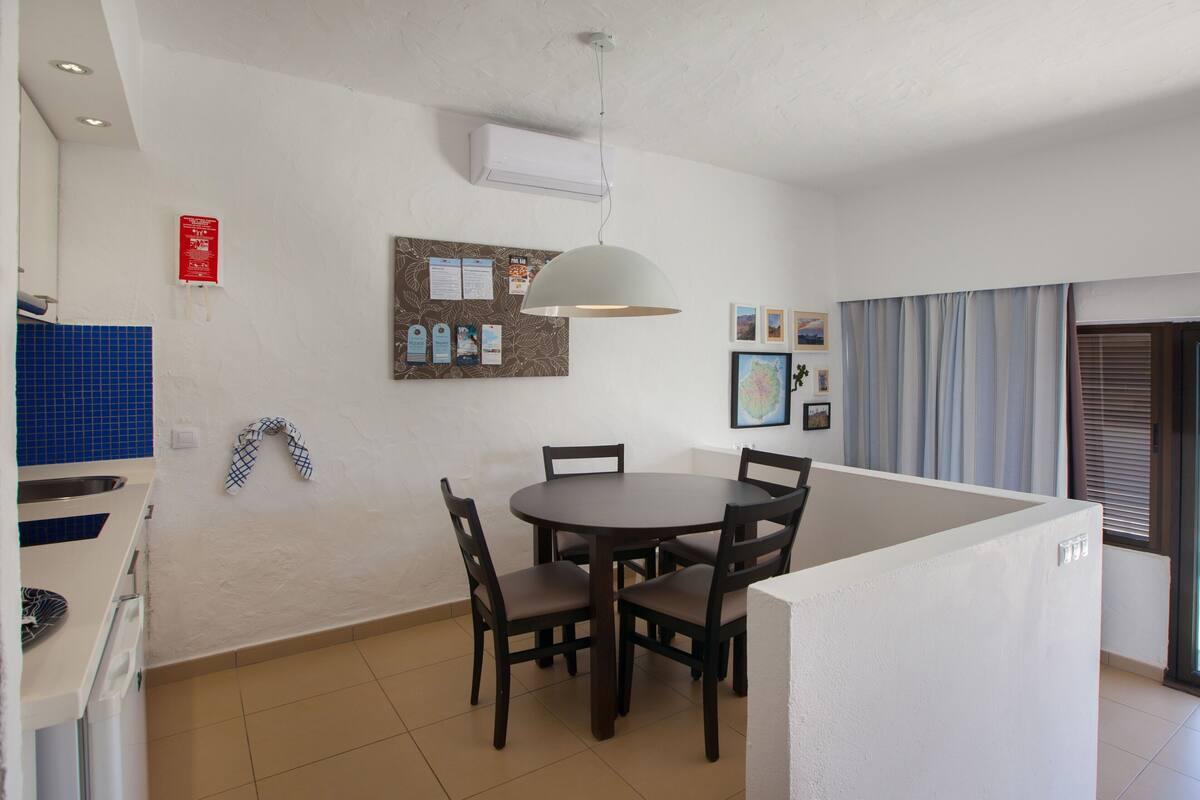 A dining area with a round table and chairs, a wall-mounted bulletin board, and a kitchen area with blue tiles.