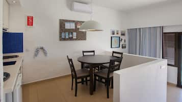 A dining area with a round table and chairs, a wall-mounted bulletin board, and a kitchen area with blue tiles.