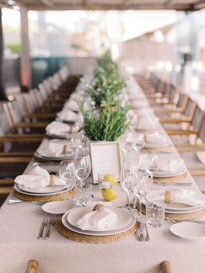 A long dining table set with white plates, gold-rimmed glasses, and silverware, with a centerpiece of greenery and a small yellow fruit.