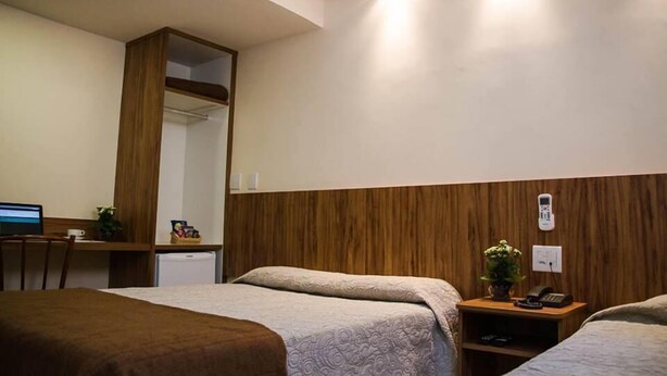 A hotel room with two beds, a desk with a computer, a small fridge, a mirror, and a bedside table with a phone and a plant. - Hotel Saint Martin (Bauru)