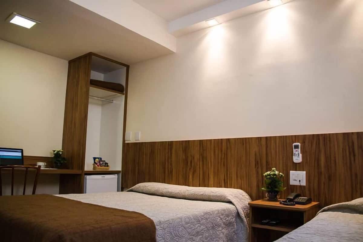 A hotel room with two beds, a desk with a computer, a small fridge, a mirror, and a bedside table with a phone and a plant.