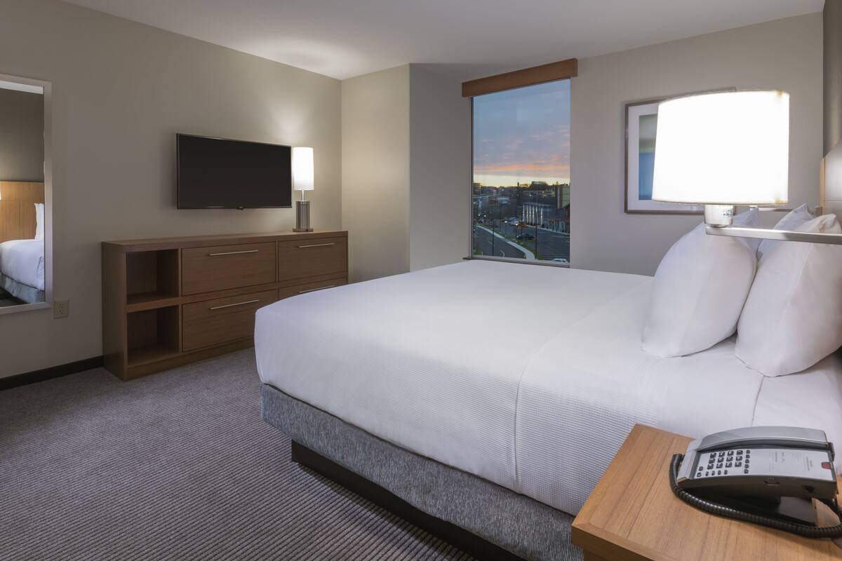 Habitación de hotel con una cama grande, un televisor de pantalla plana montado en la pared, una lámpara de noche, una mesita de noche de madera con un teléfono y una ventana que ofrece vistas a la ciudad.