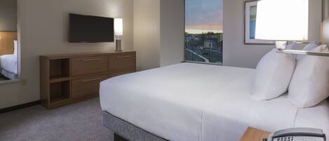 Habitación de hotel con una cama grande, un televisor de pantalla plana montado en la pared, una lámpara de noche, una mesita de noche de madera con un teléfono y una ventana que ofrece vistas a la ciudad.