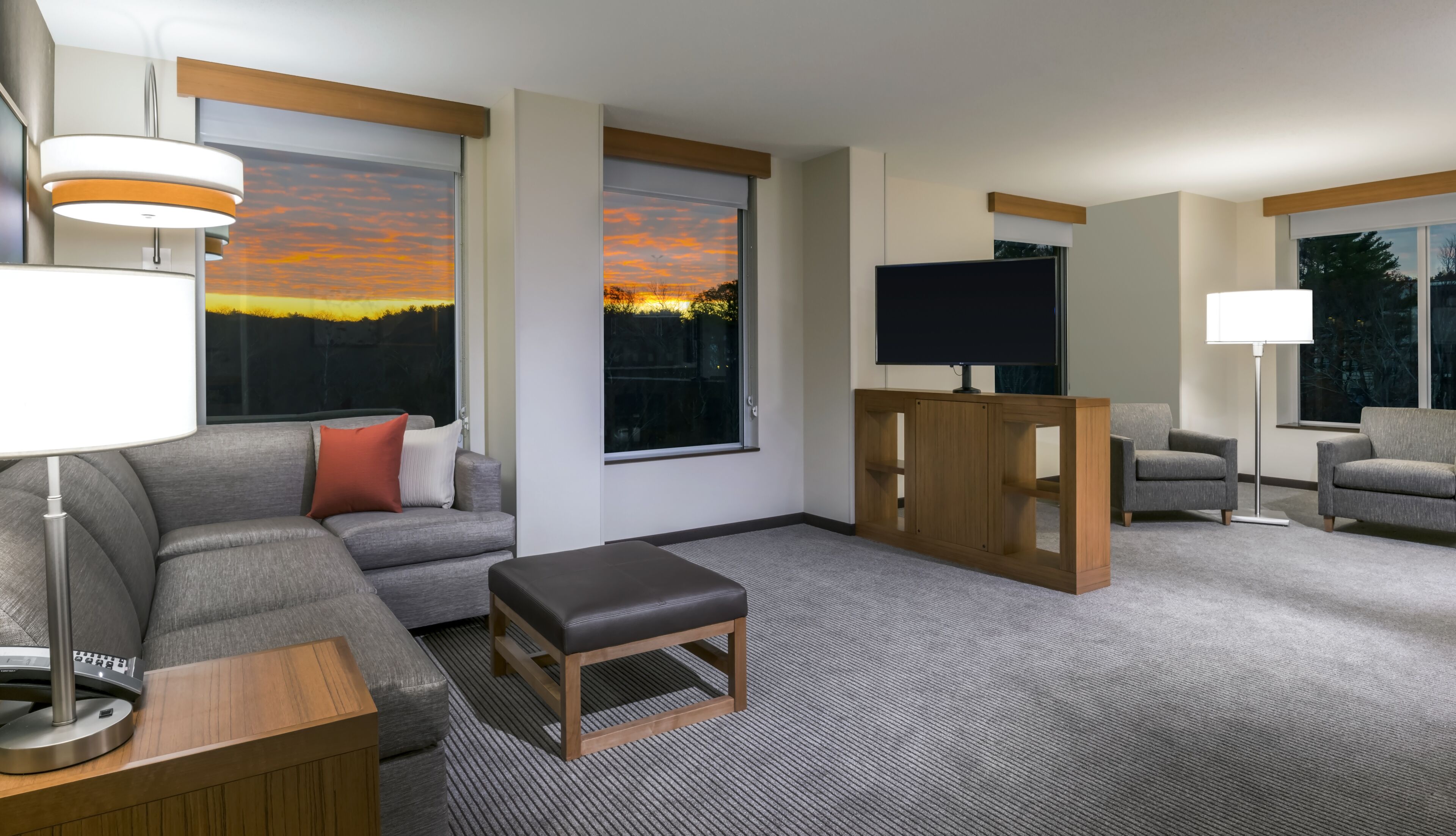 A modern hotel room with a gray sofa, a wooden bench, a flat-screen TV, and a large window with a sunset view.