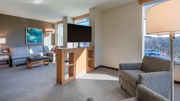 Una habitación de hotel moderna con un televisor de pantalla plana grande, un sofá gris, un mueble de entretenimiento de madera y un amplio ventanal con vistas al exterior.