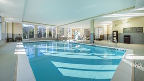 Una piscina interior con paredes de vidrio transparente, una estación de salvavidas y un área iluminada por el sol con sillas.