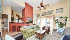 A spacious living room with a green sofa, a brown leather chair, and a glass coffee table. There is a kitchen area with red cabinets and a TV on the wall. - OCEAN WAVES HEARD FROM 2 BALCONIES! 2 min walk to Beach;3 Baths, WIFI, 3 baths (Jamaica Beach)