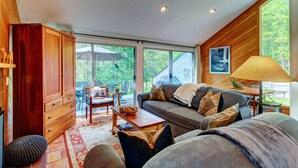 Een gezellige woonkamer met een bank, een houten kast en een eettafel. Een groot raam biedt uitzicht op het landschap.