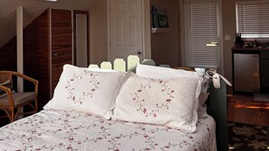 Ein Schlafzimmer mit schrägverdeckter Decke, Dachfenster, einem Bett mit floralem Bettzeug, einem Sessel und einer kleinen Küchenzeile.