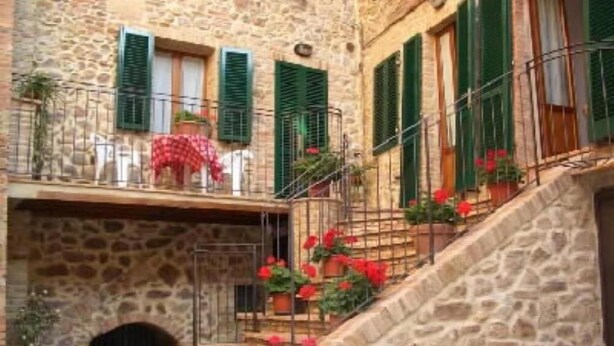 Un edificio de piedra con contraventanas verdes, un balcón con mantel a cuadros rojos y plantas en macetas.